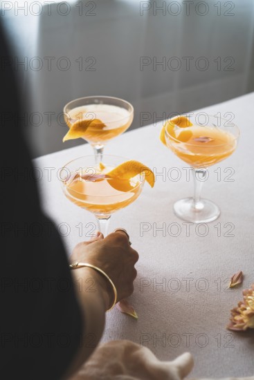 Cropped unrecognizable person holding a glass of vibrant mandarin cocktail, garnished with an orange peel, set on a sophisticated table. This image presents a perfect blend of luxury and refreshment