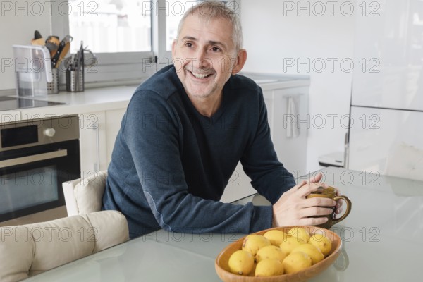 A cheerful senior man sits in a bright kitchen, holding a mug, with a basket of lemons nearby The atmosphere is cozy, highlighting a serene start to the day