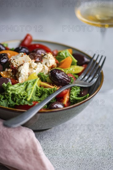 Close-up of a vibrant salad with raw kale leaves, assorted cherry tomatoes, sliced red onions, BBQ olives, and spiced feta cheese, dressed with olive oil