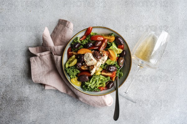 Top view of a vibrant healthy salad featuring raw kale leaves, a mix of colorful cherry tomatoes, sliced red onions, BBQ olives, and chunks of spiced feta cheese drizzled with olive oil on a textured gray background