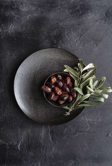Top view of a dark textured plate filled with glossy Greek BBQ olives garnished with fresh green olive leaves, presenting a rustic and appetizing look, ideal for Mediterranean cuisine themes