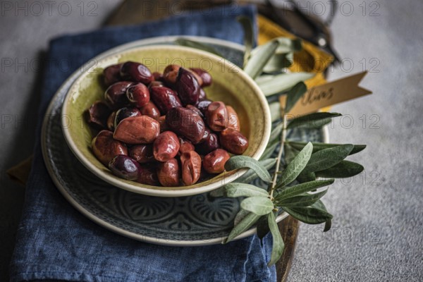 Bowl of vibrant Greek olives, accompanied by a sprig of fresh olive leaves, in a blue napkin. with a tag labeled Olives, evoking a Mediterranean vibe