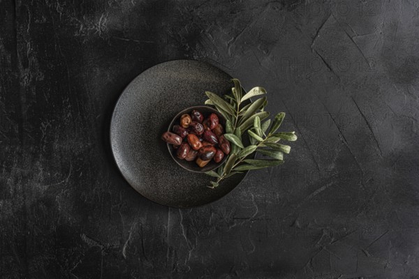 Top view of a dark textured plate filled with glossy Greek BBQ olives garnished with fresh green olive leaves, presenting a rustic and appetizing look, ideal for Mediterranean cuisine themes