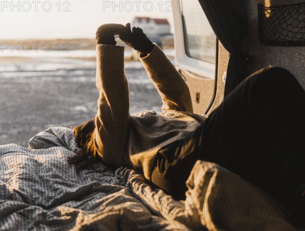 Unrecognizable woman lounging in a van during a camping trip in Iceland. She stretches her arms against the sunlight, creating a cozy, warm atmosphere