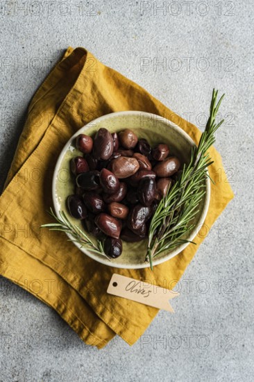 Top view of a ceramic bowl filled with Greek BBQ olives, accented with fresh rosemary sprigs, presented on a vibrant yellow napkin. Perfect for a Mediterranean-inspired snack or appetizer