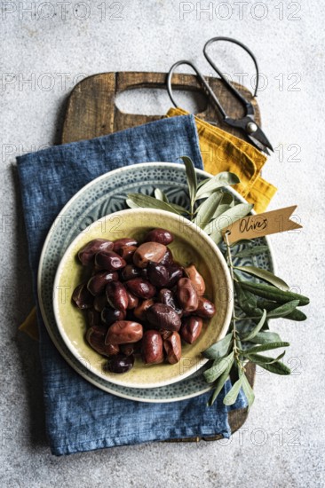 Top view of a ceramic bowl filled with Greek BBQ olives. The setting includes rustic kitchen textiles and fresh herbs, providing a homely and flavorful presentation