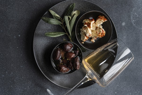 Top view of a sophisticated arrangement featuring Greek BBQ olives in a small black bowl, a portion of crumbled feta cheese seasoned with herbs, a glass of dry white wine, and fresh sage leaves on a dark, textured surface. Ideal for a gourmet Mediterranean appetizer setting