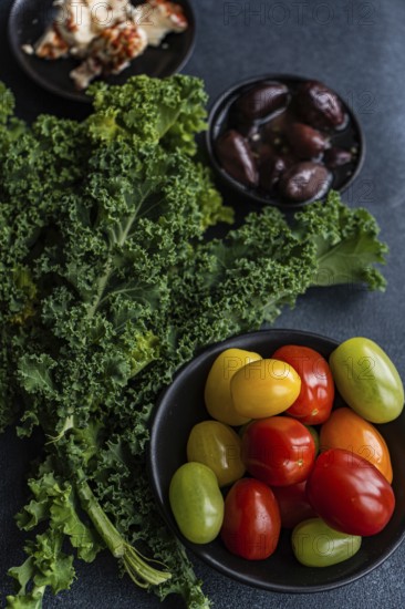 Top view of a healthy salad featuring raw kale leaves, a mix of colorful cherry tomatoes, red onion slices, BBQ-flavored olives, and spiced feta cheese, all drizzled with olive oil on a textured dark background