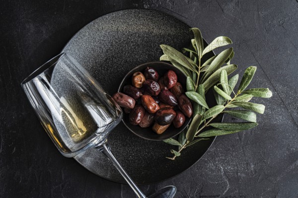 Top view of a sophisticated arrangement featuring Greek BBQ olives nestled in a black bowl with fresh bay leaves, accompanied by a glass of dry white wine, all set against a dark, textured backdrop