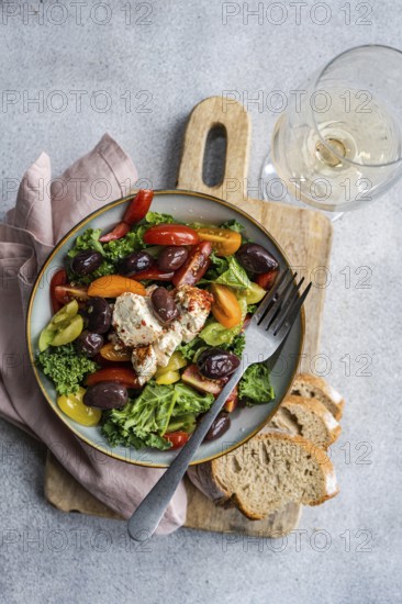Top view of a healthy salad featuring raw kale leaves, cherry tomatoes in various colors, red onion slices, olives, and spiced feta cheese. Accompanied by slices of bread and a glass of white wine on a textured grey background