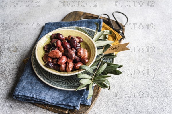 Ceramic bowl filled with Greek BBQ olives, served alongside fresh olive leaves and a rustic denim napkin, staged on a wooden chopping board
