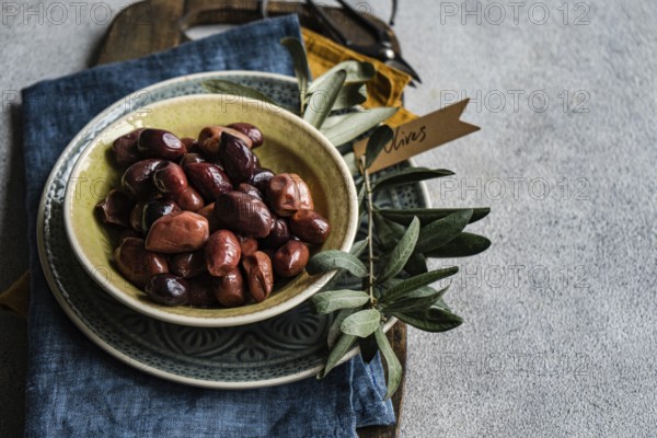 Ceramic bowl filled with Greek BBQ olives, accompanied by a strand of fresh olive leaves and a decorative tag labeled Olives. Set against a rustic backdrop with a vintage napkin, this composition epitomizes a delightful Mediterranean snack setup