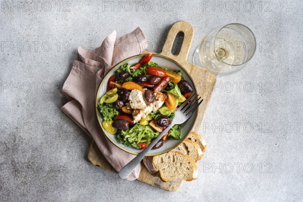 Top view of a salad featuring raw kale, assorted cherry tomatoes, red onion slices, barbecue-flavored olives, and spiced feta cheese, artfully drizzled with olive oil. The dish is complemented by slices of rustic bread and a glass of white wine