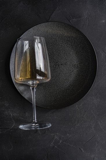 From above, an elegant glass of dry white wine placed carefully on a textured black plate, reflecting sophistication and calmness against a stark, dark background