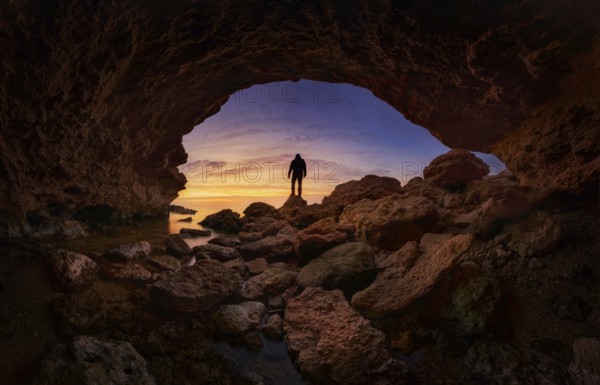 A stunning silhouette of a person standing on rocky terrain under a vibrant sunset sky, viewed from a cave at Minas Romanas, Torre de la Horadada, Alicante, Spain