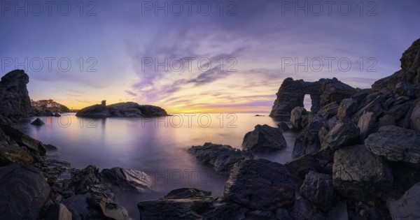 A stunning sunset at Cala Flores, Cabo de Palos, Cartagena, Murcia, featuring rugged coastal rocks, a natural stone arch, and serene waters under a vibrant, colorful sky