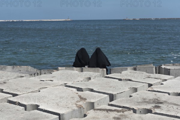 Two Muslim women wearing black abaya or chador sit on the concrete breakwater on the Mediterranean Sea front of Alexandria, Egypt