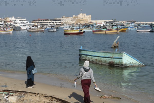 Alexandria, Egypt. December 1st 2022 Two Muslim women walk on the beach near a sunken boat in front of the citadel of Qaitbay, the city of Alexandria, the delta region of Egypt