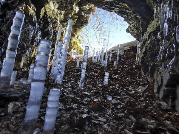 Icicles grow in a rock cave, surrounded by autumn leaves and sunlight, migrate in the Franconian Forest nature park Park