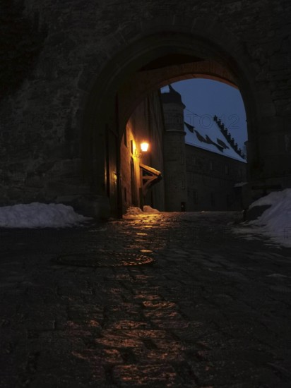 View of the courtyard of Rosenberg Fortress through a medieval archway, illuminated at night, surrounded by dark, snow-covered surroundings, Kronach, Frankenwald nature park Park