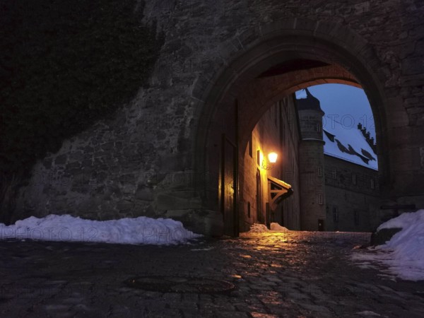 View of the courtyard of Rosenberg Fortress through a medieval archway, illuminated at night, surrounded by dark, snow-covered surroundings, Kronach, Frankenwald nature park Park