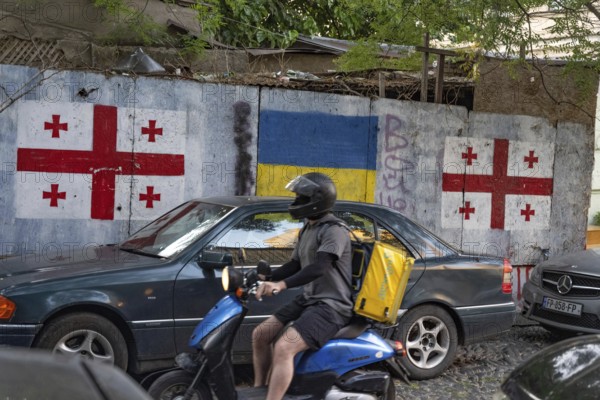 Tbilisi, Georgia. August 26th 2025. A motorcycle delivery driver rides past the flags of Georgia and Ukraine painted on the wall of a Tbilisi side street in the Georgian capital