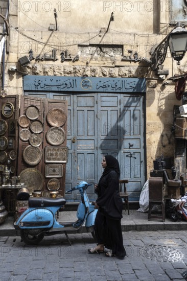 Cairo, Egypt. April 15th 2025. A Muslim woman walks past a blue motor scooter and antique shop in the Khan el Khalili Bazaar district of the ancient Islamic district of Cairo, the Egyptian capital