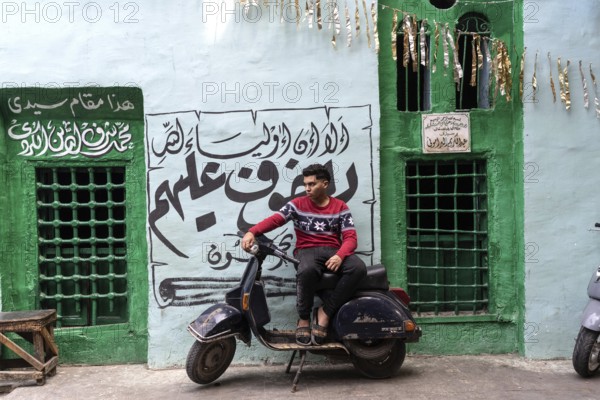 Cairo, Egypt. April 15th 2025. A young Egyptian man sits on a motor scooter outside a brightly painted tomb with Arabic inscriptions in the Islamic quarter near Midan Hussein and the Khan el Khalili Bazaar, Cairo, Egypt