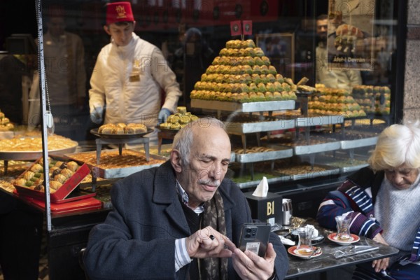 Istanbul, Turkey. January 3rd 2025. An elderly gentleman using his mobile phone outside a Turkish sweetshop in a central shopping district of Istanbul, Turkey