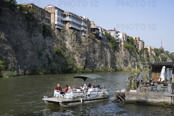 Tbilisi, Georgia. July 29th 2025. A tourist river cruise passes under building sitting precariously on a cliff above the Mtkvari or Kura River in the center of Tbilisi, Georgia