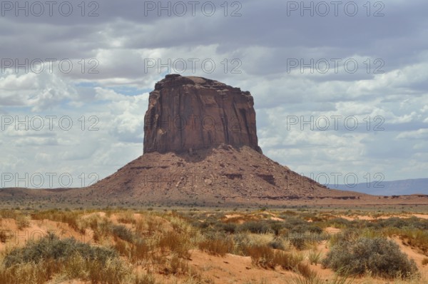 Monumental rock rises from sandy desert under cloudy sky, Monument Valley, Navajo Tribal Park, Navajo Nation Reservation, Utah, Arizona, USA