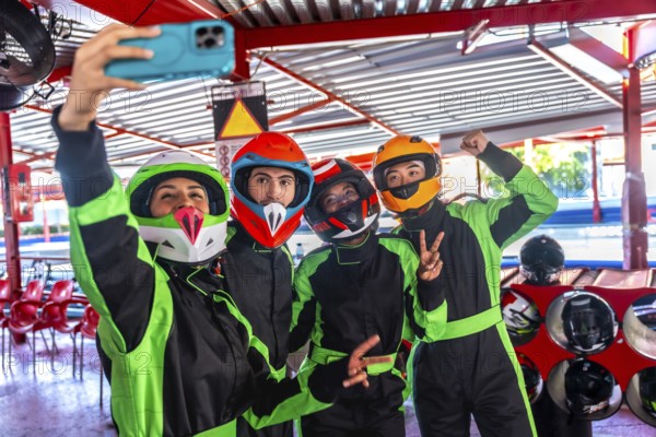 Group of four diverse friends wearing colorful helmets and racing suits, smiling and posing for a cheerful selfie at an indoor go karting track, enjoying a fun leisure activity