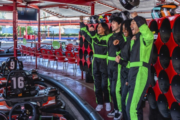 Diverse friends in racing suits celebrating with fists pumped up, feeling excited and happy after completing a thrilling go kart race at an indoor track
