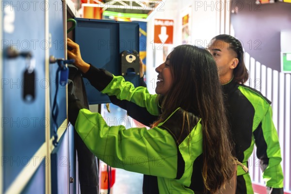 Young woman smiling while reaching into a locker to retrieve gear, with a man standing beside her, both wearing green and black racing suits, preparing for the karting experience