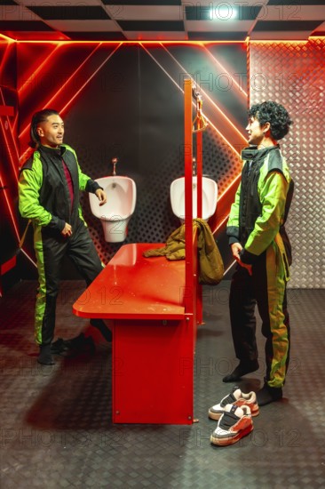 Two young men in green and black jumpsuits chat in a red neon locker room, gearing up and bonding before an adrenaline filled go kart race