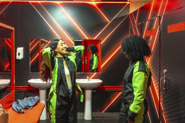 Two young women in neon lit locker room laughing and suiting up in racing jumpsuits, bonding before an energetic indoor go karting session with friends and fast paced fun