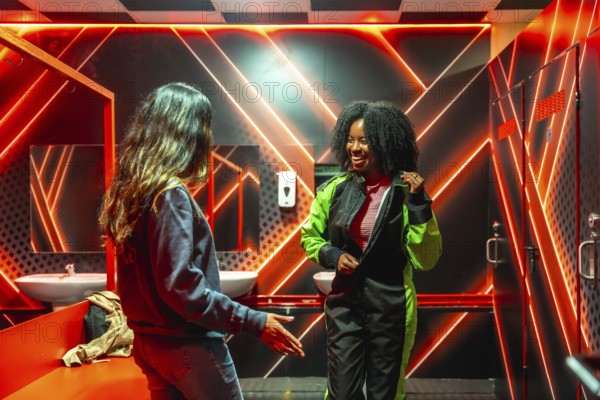 Two cheerful women are preparing for a gokarting race in a neon lit locker room, with one friend putting on her brightly colored racing suit while laughing