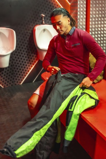 Young man sitting in a changing room, putting on a black and green racing suit for an upcoming karting session, focusing on preparing for the recreational sport challenge