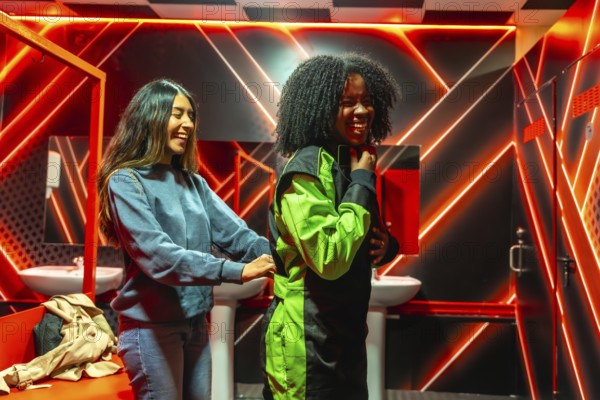 Two diverse friends laughing and helping each other put on racing suits in a changing room with vibrant neon lights, anticipating a fun go kart race activity