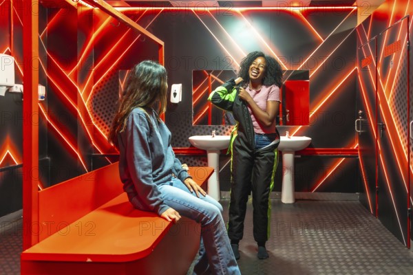 Two diverse young women laughing and dressing in race suits inside a modern changing room with red neon lights, preparing for a thrilling karting ride