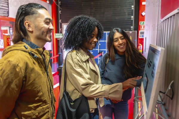 Diverse group of young adult friends laughing and talking while interacting with a self service touchscreen for an activity, suggesting leisure, technology, and friendship concepts