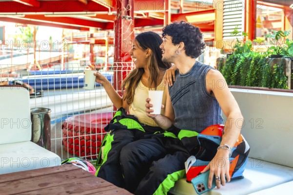 Young happy couple laughing and enjoying drinks, wearing racing suits and holding helmets after an exciting go karting race. They are relaxing together, sharing memories of the fun experience
