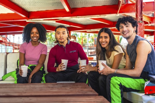 Multiethnic group of young adults in racing suits relaxing at a table with drinks, smiling and laughing together after karting for a fun, social leisure moment at the track