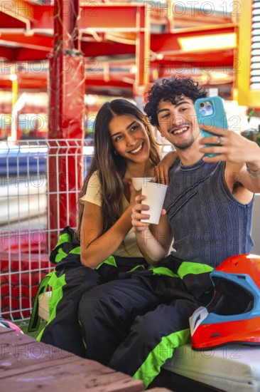 Young couple wearing racing suits taking a selfie during a karting break, showing happiness and connection as they enjoy a recreational activity together