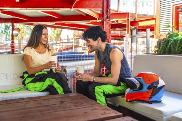 Young couple in racing suits sharing coffee and laughter at a trackside table after karting, relaxed conversation and bonding during a fun outdoor weekend activity