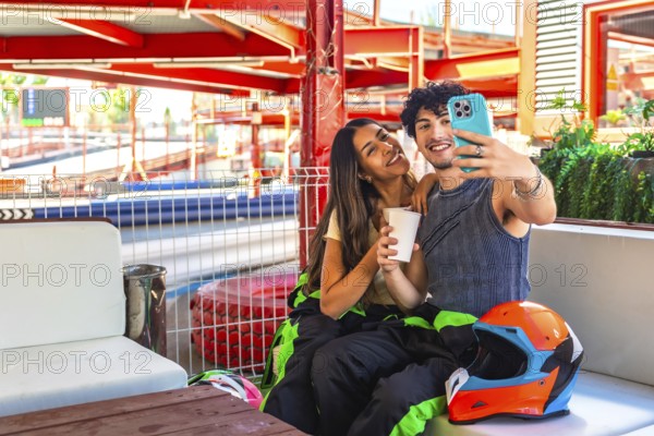 Young couple wearing racing suits sitting on a bench at a go kart circuit, smiling and taking a selfie with a smartphone, celebrating a fun racing date