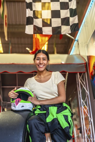 Young woman in racing suit sits in a go kart holding her helmet beneath a checkered flag, smiling confidently before a fun, competitive race on an indoor track