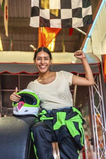 Young woman smiling and raising a fist in triumph, holding a karting helmet, wearing a racing suit, and standing under a checkered flag, symbolizing achievement and success in sports