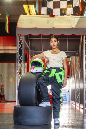 Woman wearing a racing suit and helmet standing confidently by tires and checkered flags at an indoor karting track, ready for leisure or sport activity