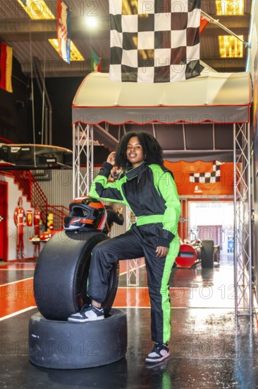Young african american woman posing confidently in a neon green and black racing suit and helmet, standing next to tires at an indoor go kart track with a checkered flag visible